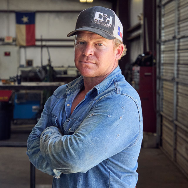 A man, Wes Stevenson, in a worn denim shirt and baseball cap stands with arms crossed in a metal workshop.